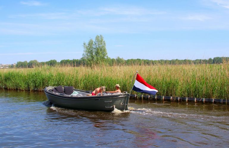 Sloep met twee personen en Nederlandse vlag vaart langs een rietkraag