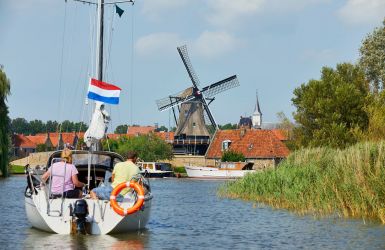 Zeilboot vaart richting een dorp met molen, omringd door water en riet